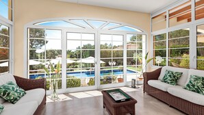 A sunlit room with a large glass door leading to a pool, wicker furniture, and a view of greenery.