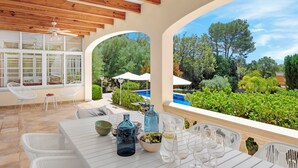 A patio with a table set for dining, white chairs, and a view of a swimming pool and greenery.