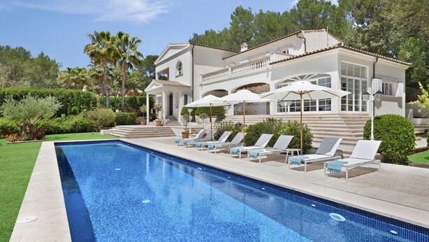 A swimming pool with lounge chairs and umbrellas, surrounded by a well-maintained garden and a large building with a balcony.