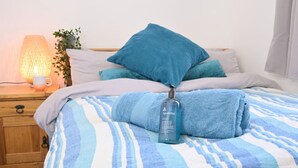 A neatly made bed with a striped blue and white bedspread, a blue bottle of Apothicie hydrating hand wash, and a wooden nightstand with a lamp and a potted plant.