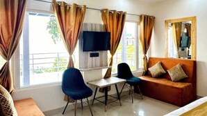 Habitación de hotel con cama, sofá, escritorio y dos sillas. Tiene un televisor en la pared y una ventana con cortinas.