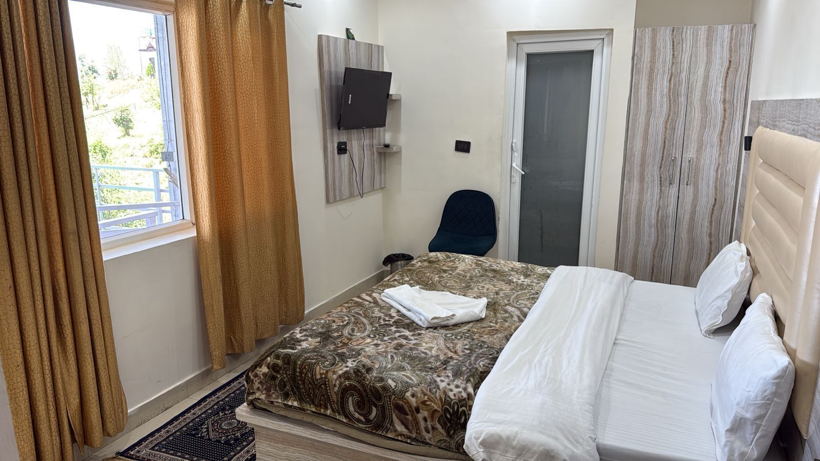 A hotel room with a bed, a TV mounted on the wall, a door, and a window with curtains.