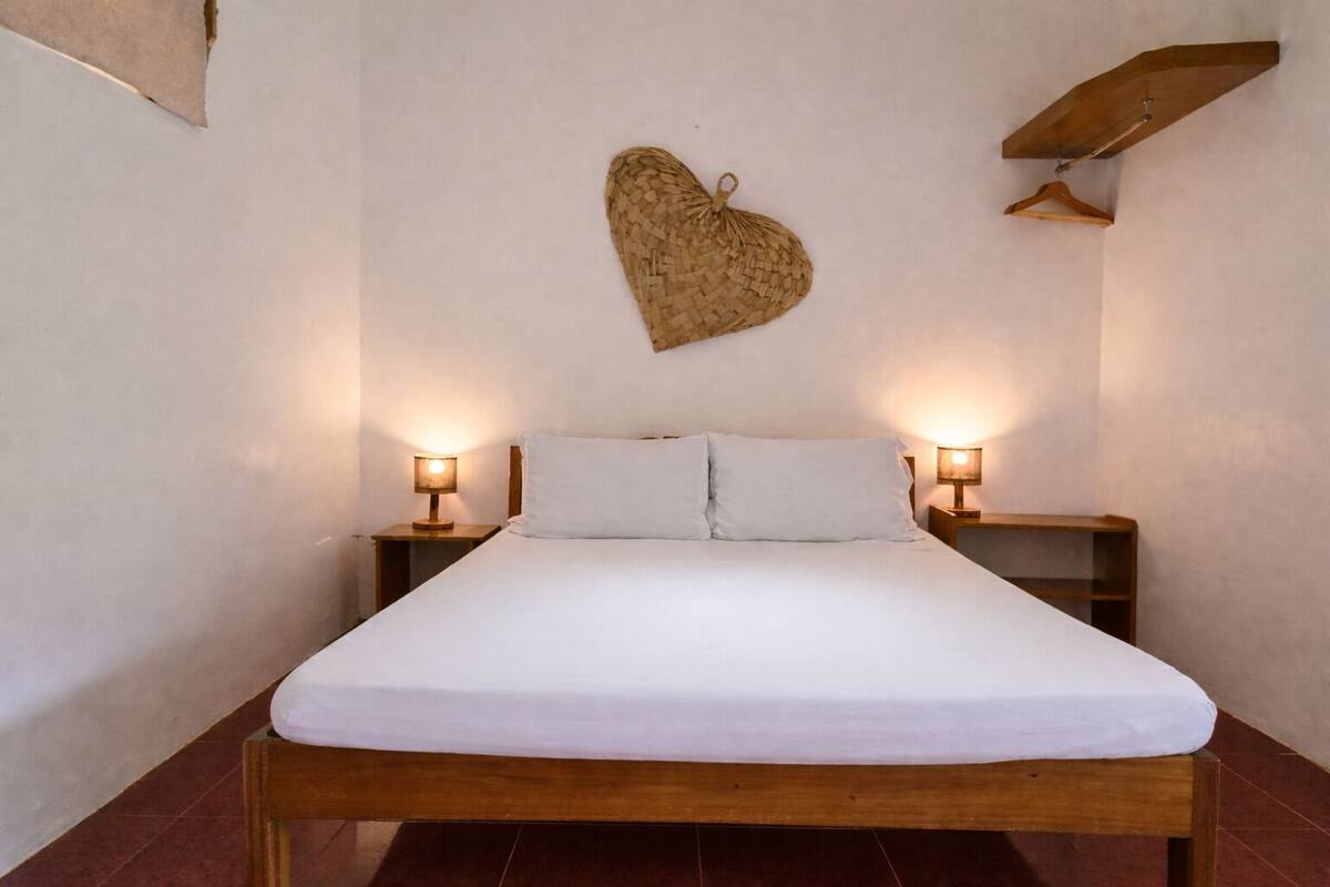 Un lit simple avec une literie blanche, deux tables de chevet avec des lampes, une tête de lit en bois et une décoration murale tissée.