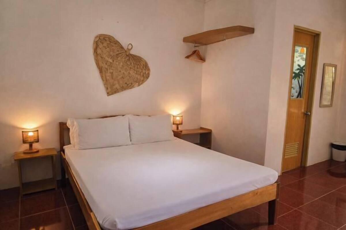 Un lit simple avec un cadre en bois, deux lampes de chevet, une décoration en forme de cœur fixée au mur, une porte avec une fenêtre et une petite poubelle.
