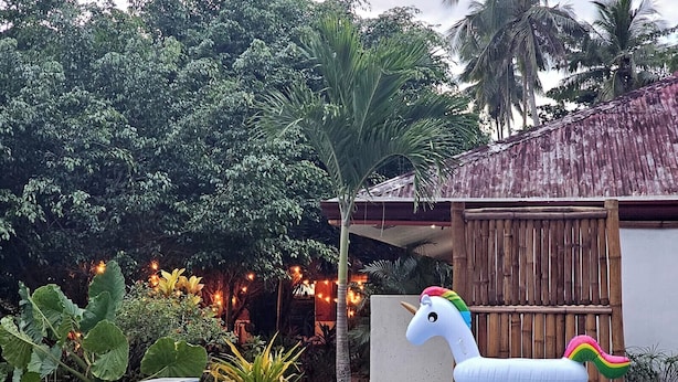 A swimming pool with a unicorn float, surrounded by lush greenery and a rustic building. - Masayana Hotel (El Nido)