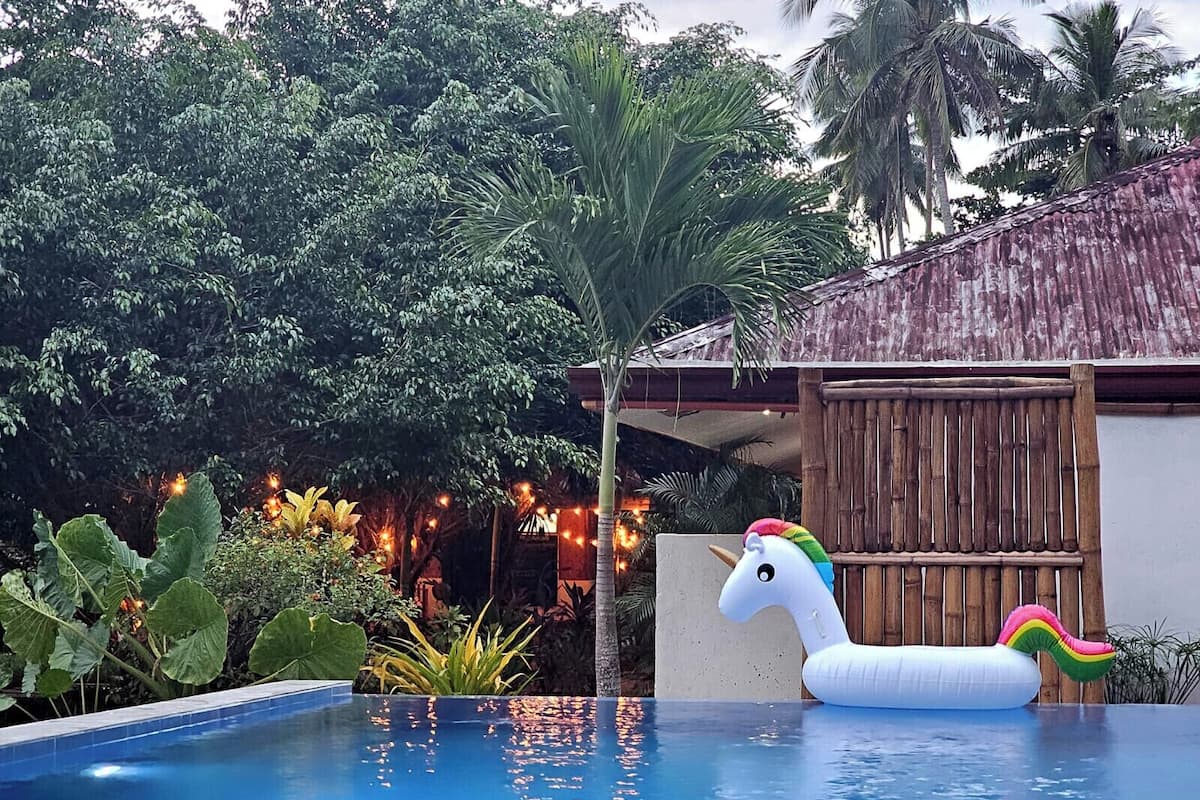 Une piscine avec un flotteur en forme de licorne, entourée d’une végétation luxuriante et bordée d’un bâtiment au style rustique.