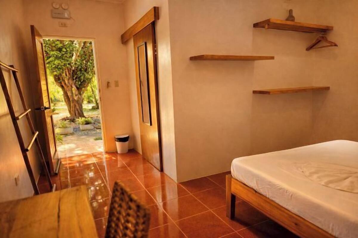 Une chambre à coucher avec un lit, une table en bois, une chaise et une vue sur un arbre par une porte ouverte.