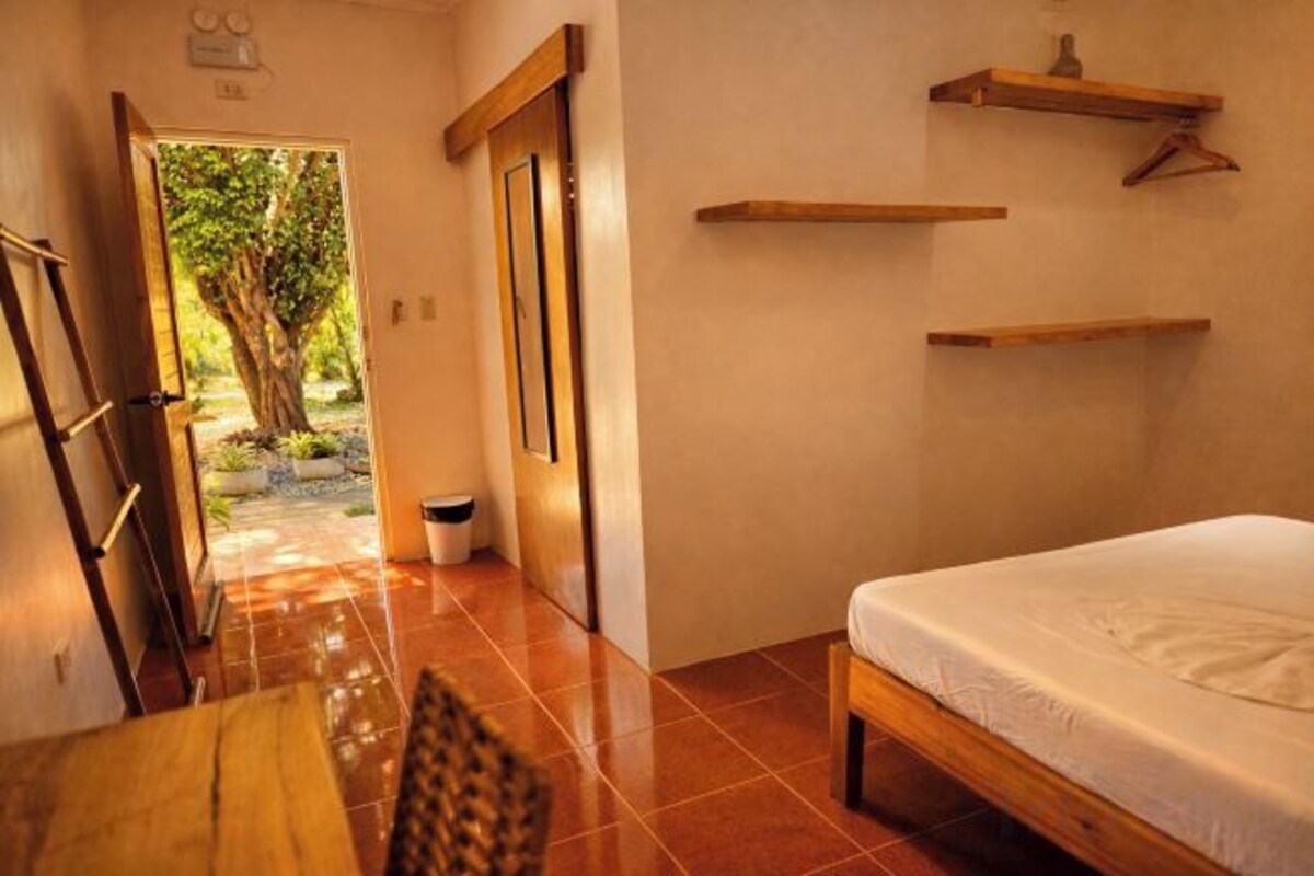 Una habitación con una cama, una mesa y una silla. Se ve una puerta que da a un espacio exterior con un árbol.