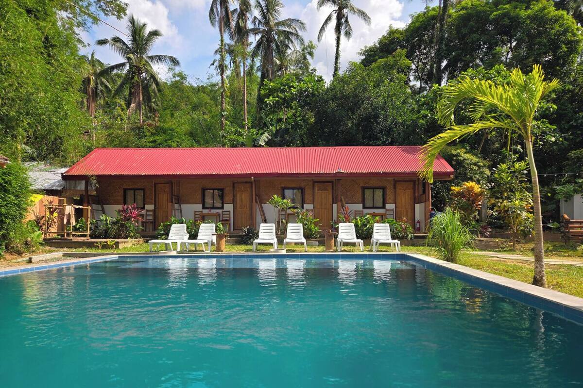 Une piscine bordée de chaises longues blanches, un bâtiment au toit rouge et une végétation tropicale.