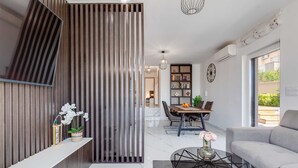 A modern living room with a sofa, a dining area with a table and chairs, a bookshelf, and a decorative plant. - Summer Dream Apartment with garden, patio and pool (Sv. Filip i Jakov)