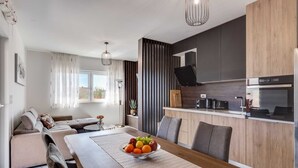 A modern kitchen with a dining area, featuring a wooden table with a fruit bowl, grey upholstered chairs, and a built-in oven. - Summer Dream Apartment with garden, patio and pool (Sv. Filip i Jakov)