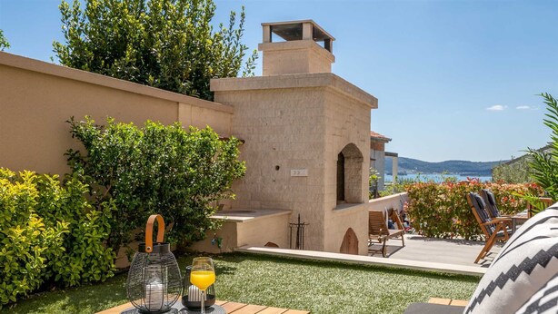 A patio with a wooden table, a book, and a glass of orange juice. There are chairs and a view of the sea in the background. - Summer Dream Apartment with garden, patio and pool (Sv. Filip i Jakov)