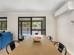A dining area with a wooden table, chairs, a bowl, and a view of a swimming pool through glass doors. - Nerita Nook (Nelly Bay)