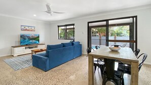 A living room with a blue sofa, a wooden dining table with chairs, a TV on a white cabinet, and a large window with a view of greenery. - Nerita Nook (Nelly Bay)