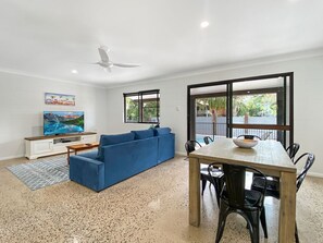A living room with a blue sofa, a wooden dining table with chairs, a TV on a white cabinet, and a large window with a view of greenery. - Nerita Nook (Nelly Bay)