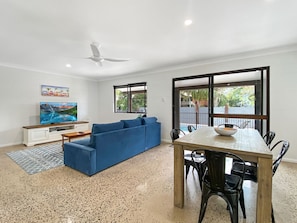 Uma sala de estar com um sofá azul, uma mesa de jantar de madeira com cadeiras, uma TV sobre um armário branco e uma janela grande com vista para o verde.