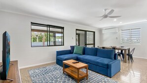 A living room with a blue sofa, a wooden coffee table, a TV on a wooden stand, a patterned rug, a ceiling fan, and a dining area with a table and chairs. - Nerita Nook (Nelly Bay)
