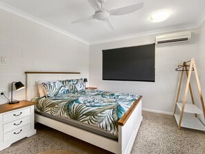 A bedroom with a bed, a nightstand, a desk, a ceiling fan, an air conditioning unit, and a TV mounted on the wall. - Nerita Nook (Nelly Bay)