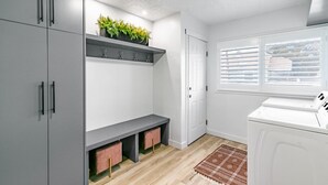 Une buanderie équipée d’un lave-linge, de meubles de rangement gris, d’un banc et d’une fenêtre avec des stores.