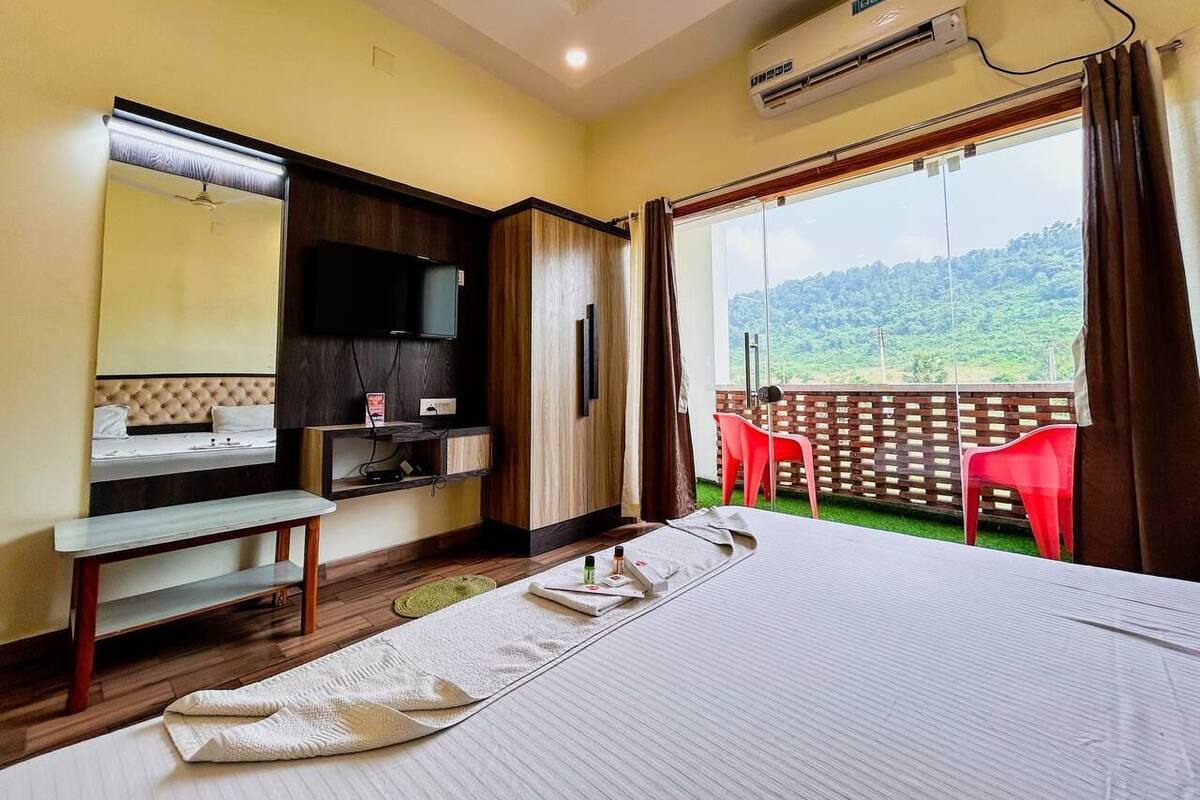 Quarto de hotel com cama, escrivaninha, televisão e varanda com vista para as colinas.