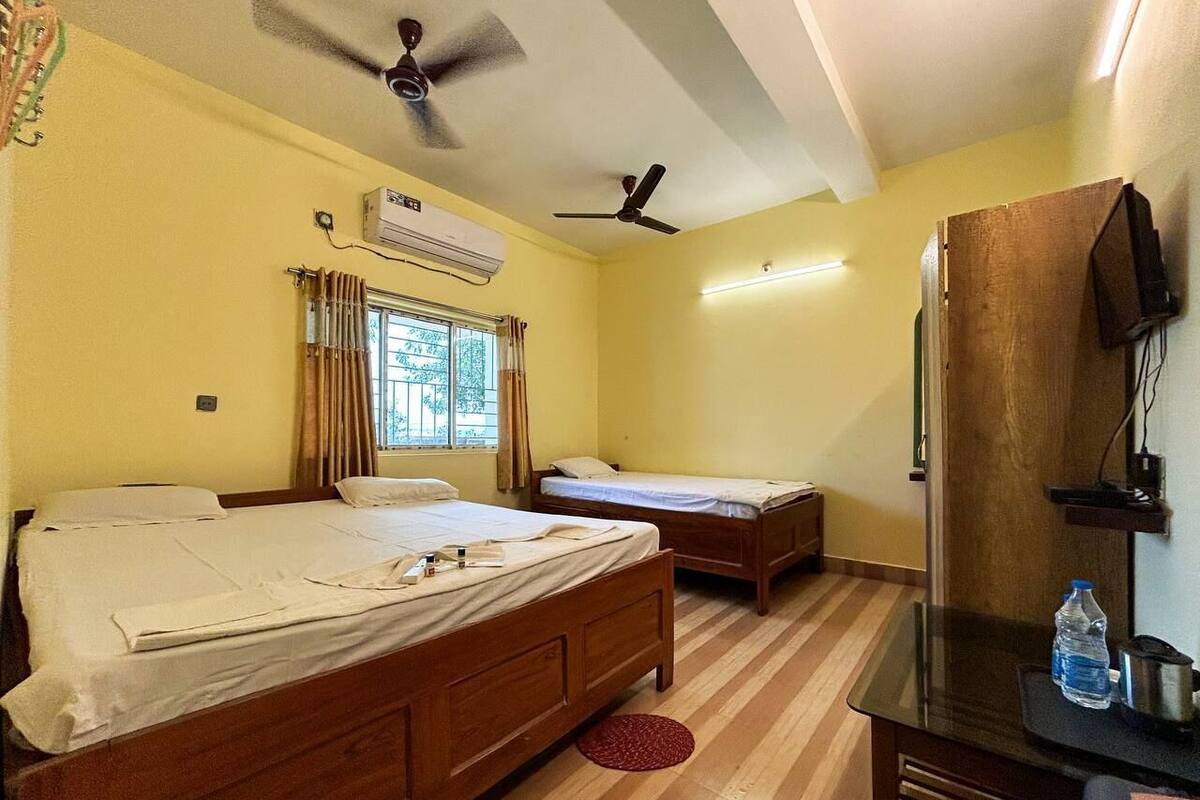 Quarto com duas camas de madeira, um ventilador de teto, ar condicionado, uma TV fixada em um armário de madeira e uma janela com cortinas.