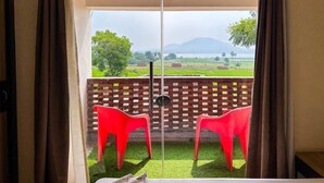 A room with a sliding glass door leading to a balcony with two red chairs and a view of a green landscape with trees and mountains. - Allure De Baranti (Raghunathpur)