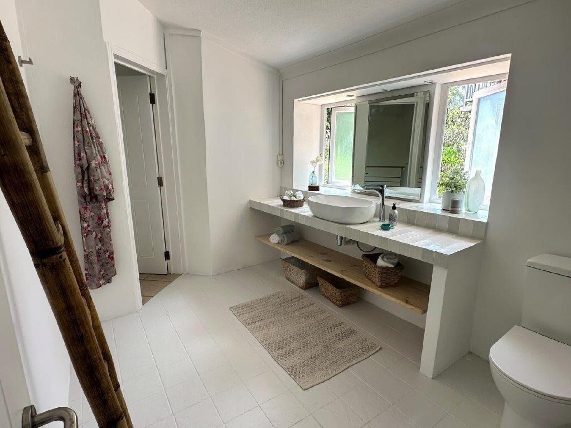 A modern bathroom with a large sink, a mirror, and a window. There is a door leading to another room and a wooden bath mat on the floor.