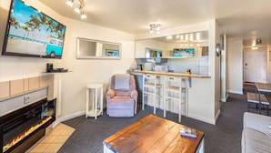 A modern living room with a fireplace, a TV mounted on the wall, a comfortable armchair, a wooden coffee table, and a kitchen area with a microwave and bar stools. - So Very Nice in Ocean Shores (Ocean Shores)