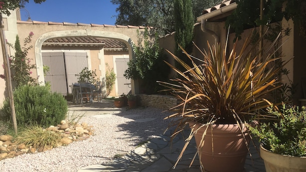 A courtyard with a stone pathway leading to a building with a tiled roof and a large arched doorway. There are potted plants and a variety of greenery around the area. - A Different Side of Provence (Jonquerettes)