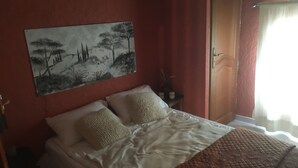 A bedroom with a bed, pillows, a painting on the wall, a nightstand, and a window with curtains. - A Different Side of Provence (Jonquerettes)