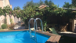 A swimming pool with a wooden deck and metal ladders, surrounded by greenery and a building. - A Different Side of Provence (Jonquerettes)
