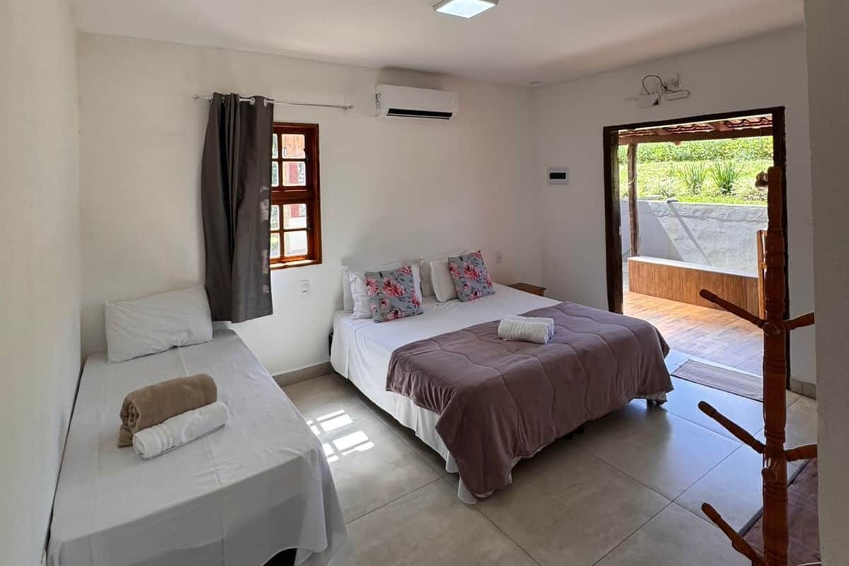 A room with two beds, a window with curtains, an air conditioning unit, and a view of greenery outside.