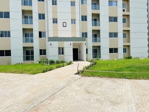 A multi-story building with a central entrance, surrounded by a paved walkway and a grassy area. - Serenity Haven 2 BR | Gated | Pool | WiFi | AC Netflix  (Sakumono)