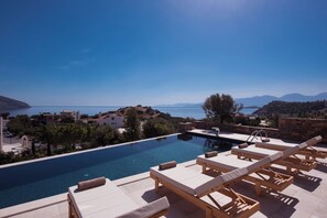 A pool area with lounge chairs and a view of the sea and hills.