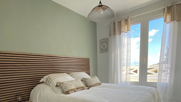A bedroom with a large bed, a wooden headboard, a bedside table with a lamp, and a view through the window. - Villa with communal pool in St palais sur mer (Saint-Palais-sur-Mer)