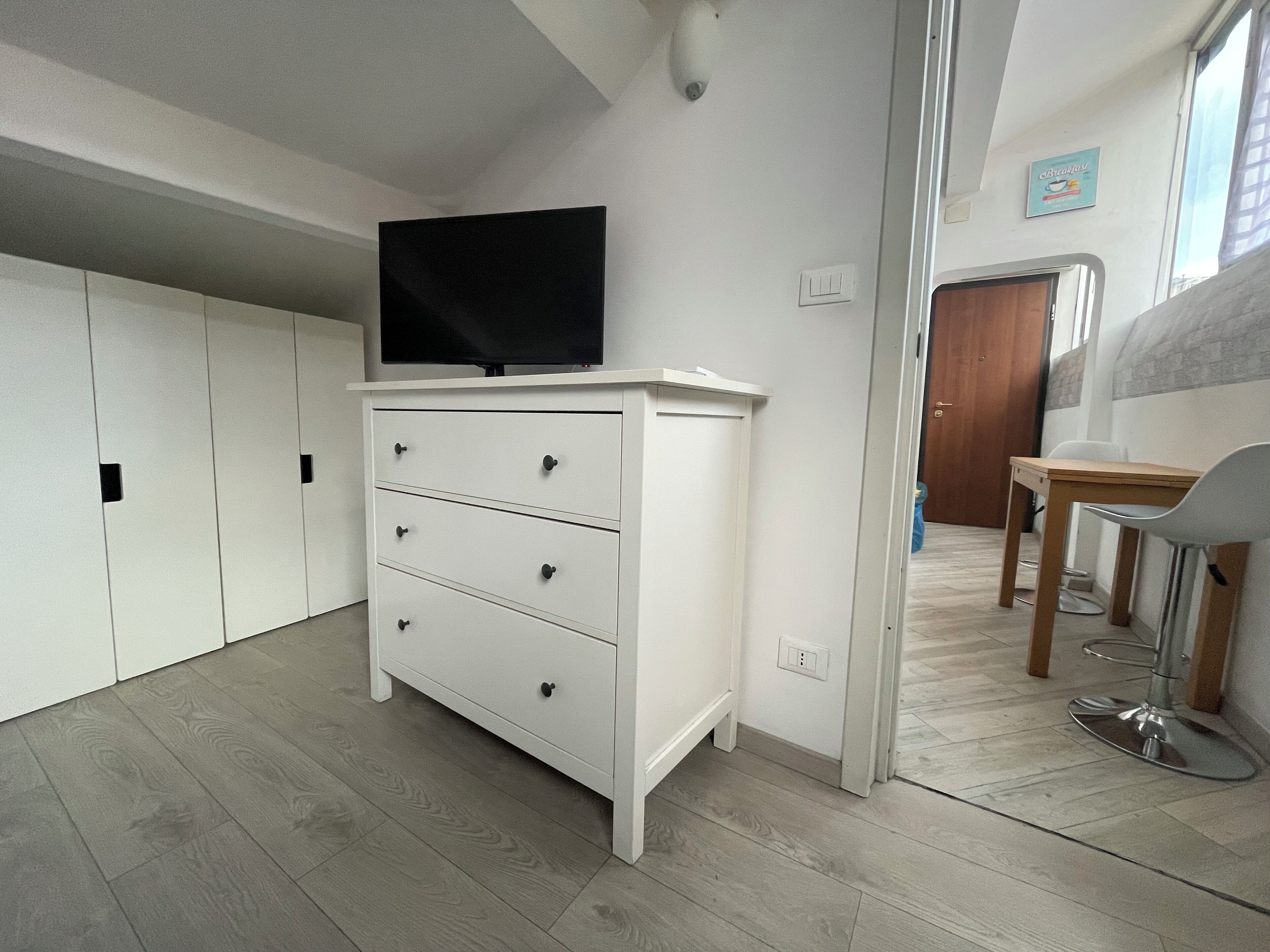 Una habitación moderna y bien iluminada con un tocador blanco, un televisor de pantalla plana y una zona de comedor con una mesa y sillas.