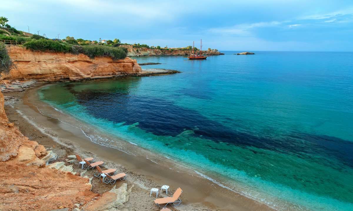 A coastal beach with clear waters, a rocky shoreline, and a boat in the distance.