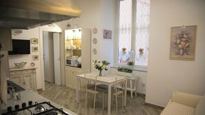 A kitchen with a stove, dining table, and chairs. There is a window with lace curtains and a painting on the wall.