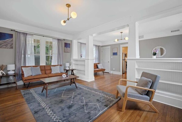 A spacious living room with a sofa, armchair, and coffee table, wooden flooring, and a large window with curtains.