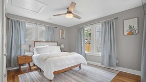 Un dormitorio con cama, mesita de noche, ventilador de techo y ventanas grandes con cortinas.
