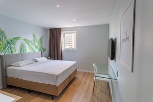 Un dormitorio moderno con cama, un escritorio de vidrio, una ventana con cortinas y un televisor montado en la pared.