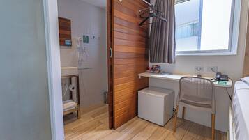 A compact hotel room with a wooden wall, a small desk with a chair, a bed, a shower area, and a window with curtains.