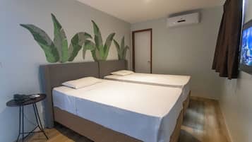 A hotel room with two single beds, a small table with a phone and remote control, and a wall-mounted air conditioner.