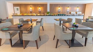 Un comedor moderno con varias mesas y sillas, un mostrador de buffet con diversos alimentos, e iluminación en la pared.