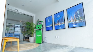 Un moderno vestíbulo de hotel con un mostrador de recepción, un quiosco verde de autoservicio y tres cuadros con vistas urbanas en la pared.