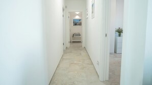 A narrow hallway with white walls and a tiled floor leading to a room with a bed and a window.