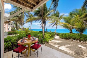 A covered outdoor dining area with a table set for two, surrounded by lush greenery and palm trees, overlooking a beach.
