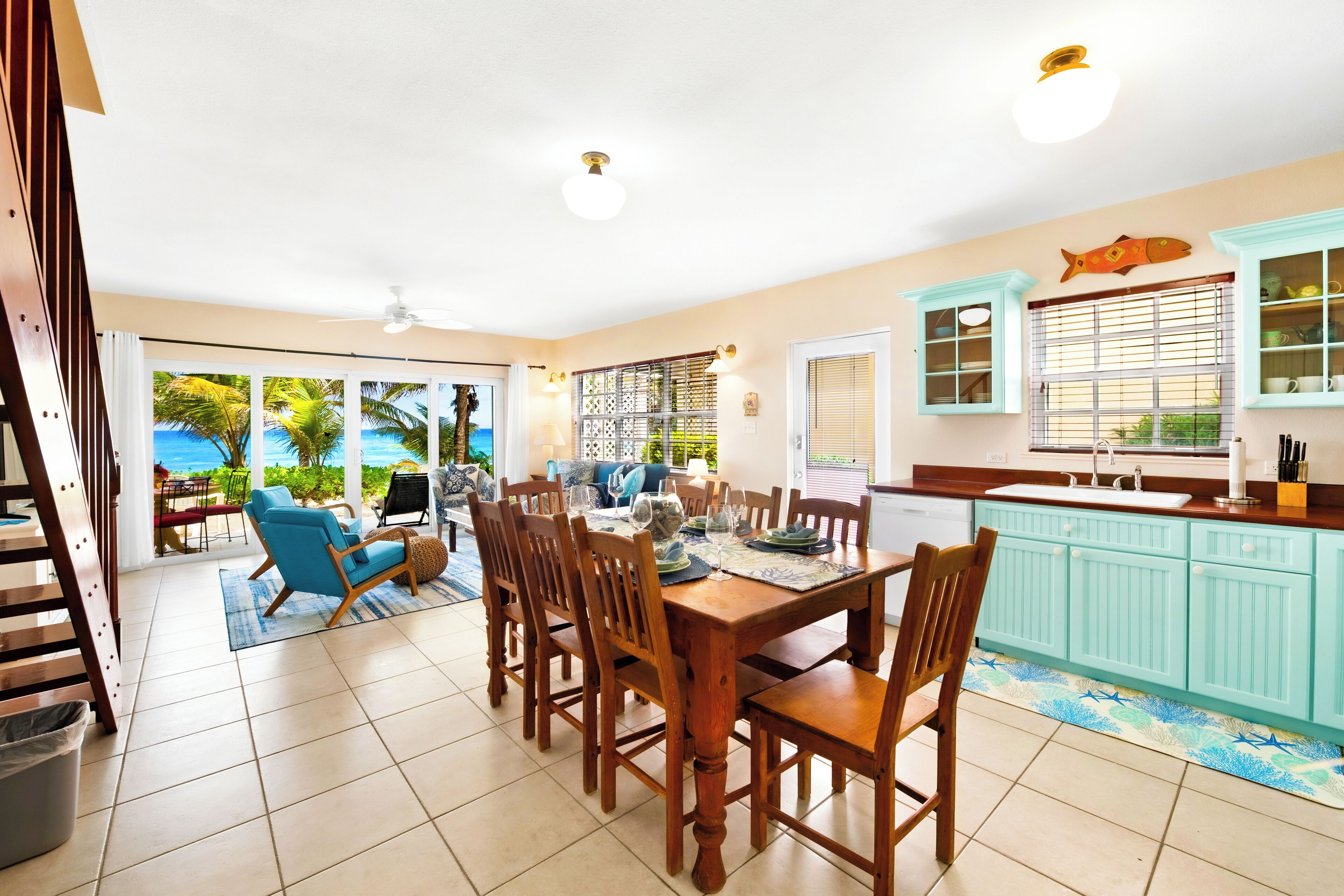 A spacious dining area with a wooden table and chairs, a kitchen with light blue cabinets, and a view of the ocean through large windows.