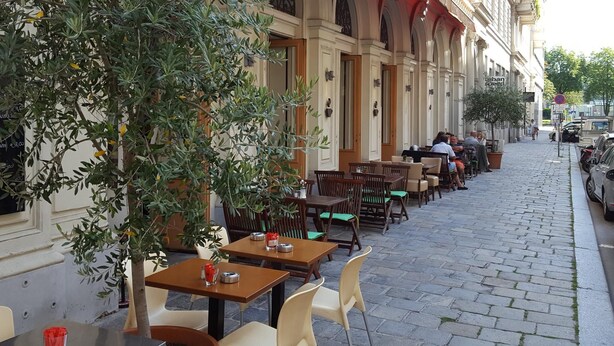 An outdoor café with tables and chairs, a tree, and a building with arched windows in the background. - Central family Apartment A16 with AC (Vienna)