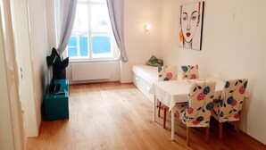 Ein Zimmer mit Holzboden, einem weißen Tisch mit floralen Stühlen, einem Bett mit einem Blumenkissen und einem Gemälde an der Wand.